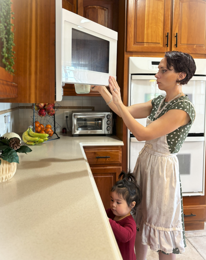 A woman in a cream colored apron opening the microwave in her kitchen. She has a toddler standing underfoot and she is placing a measuring cup of milk in the microwave.