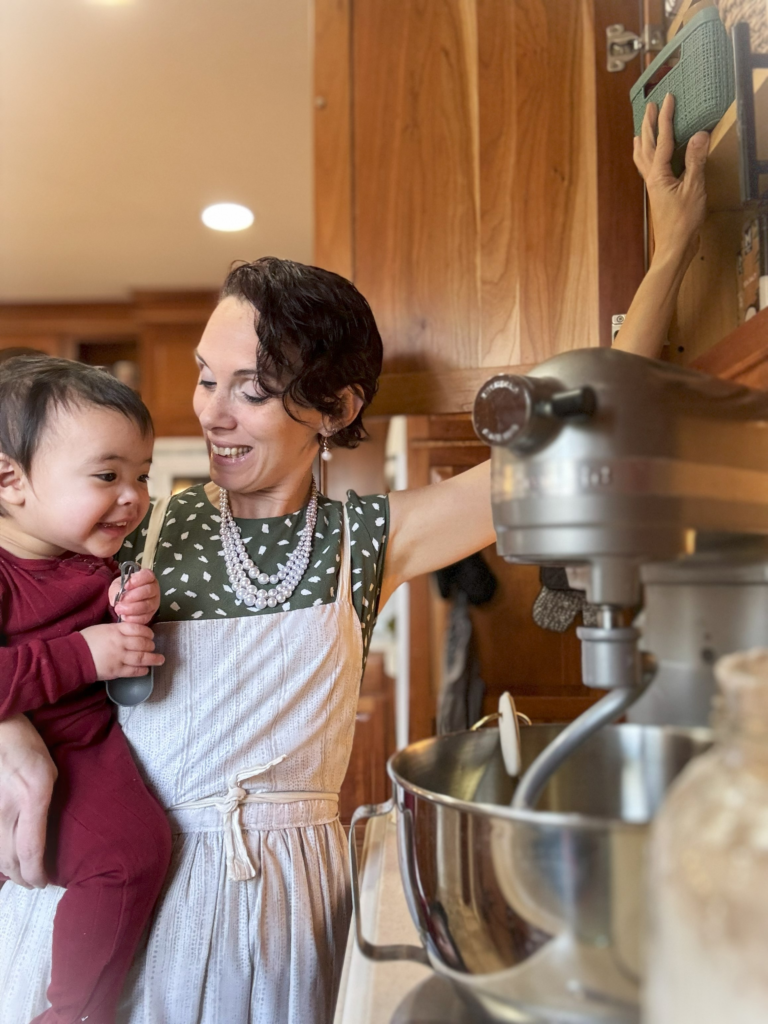 A mother is holding her baby on her hip with one hand, while the other hand is placing a basket of spices into a kitchen cabinet. There is a kitchen stand mixer in view